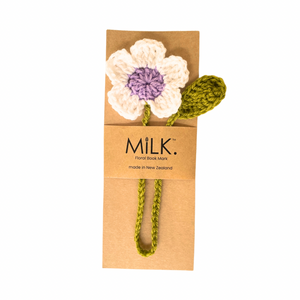 A handmade crochet flower hair clip by the brand Milk, featuring a white flower with a purple center and an olive green stem and leaf, attached to a brown kraft paper band with the brand logo.