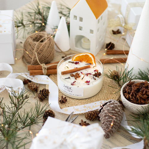 Candle with decorative elements on a table, including pine cones and cinnamon sticks, with a 'Milk' label visible.