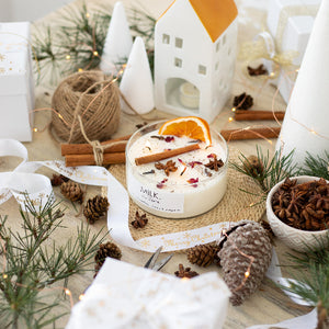 Candle with decorative elements on a table, including pine cones and cinnamon sticks, with a 'Milk' label visible.